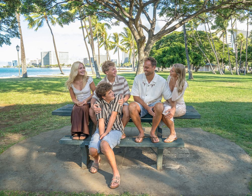 A group of people sit together on a sandy beach under palm trees during a sunny tropical day.