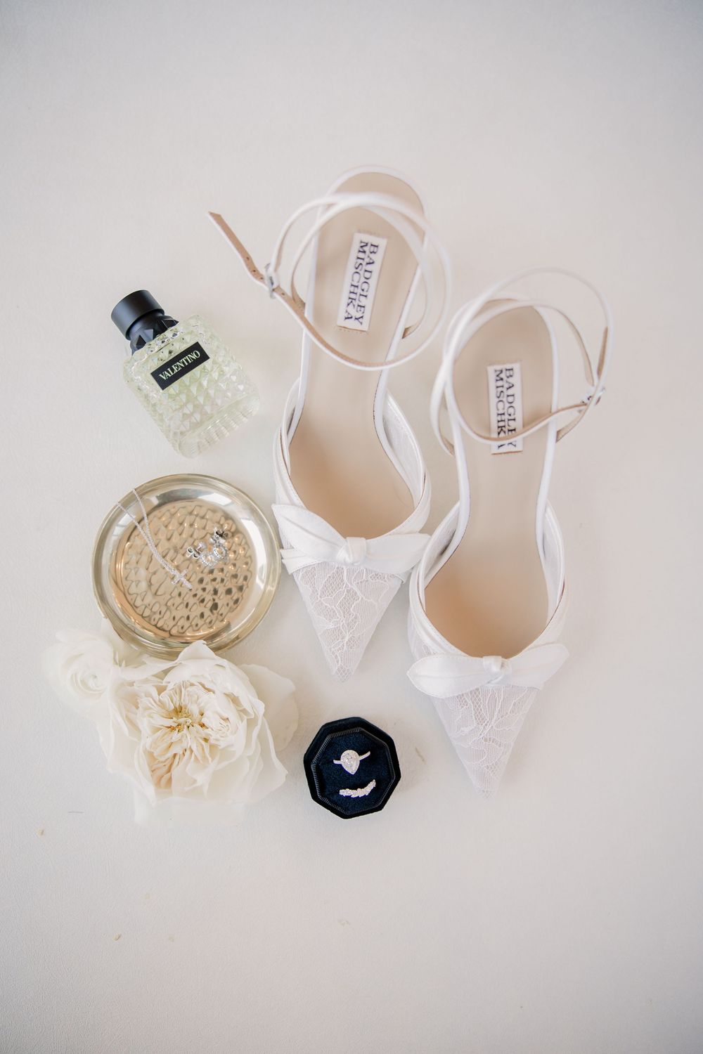 Badgley Mischka white lace bridal heels displayed with perfume bottles and floral accessories on white surface.