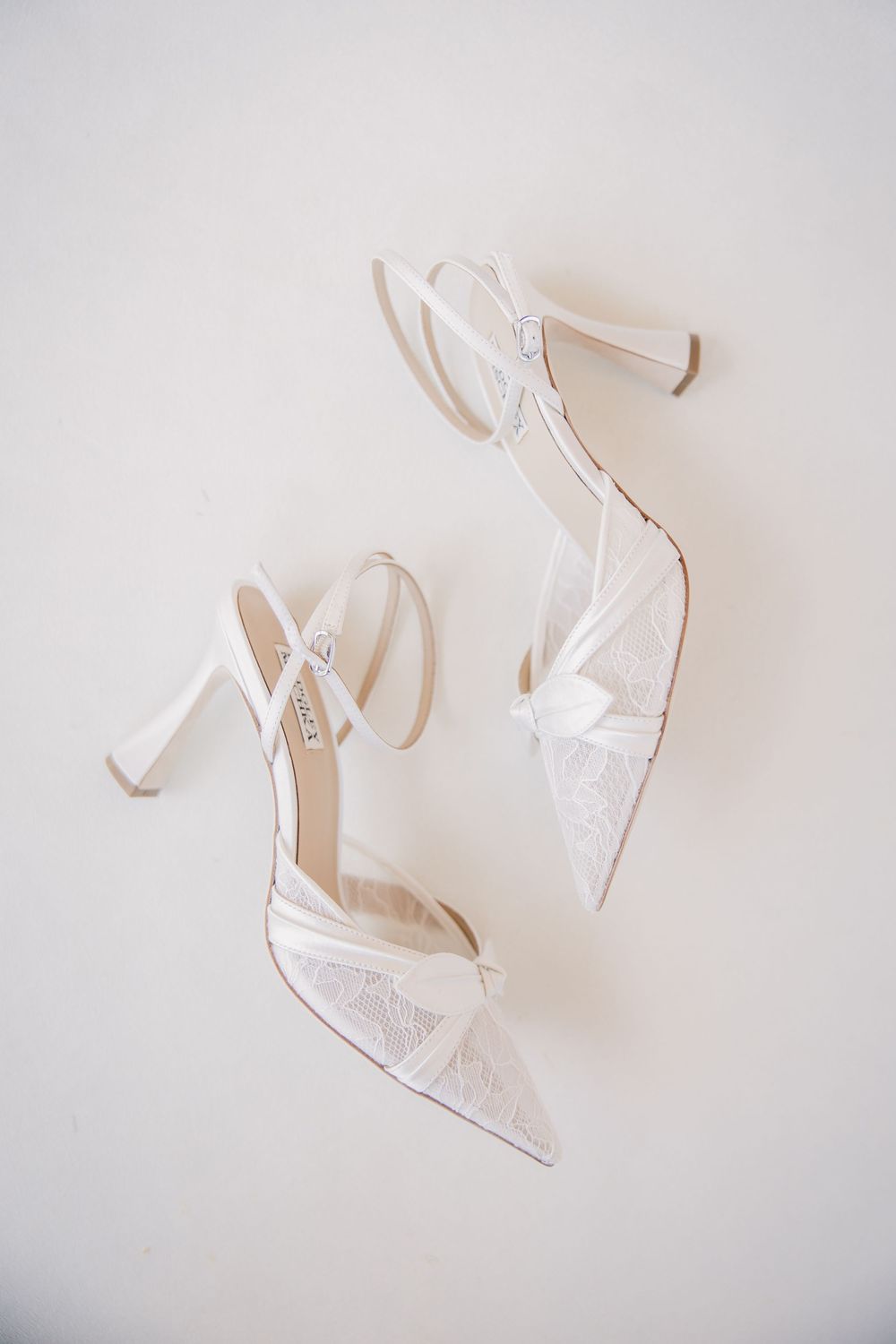 Pair of white lace pointed-toe bridal heels with ankle straps laid flat on a white surface.