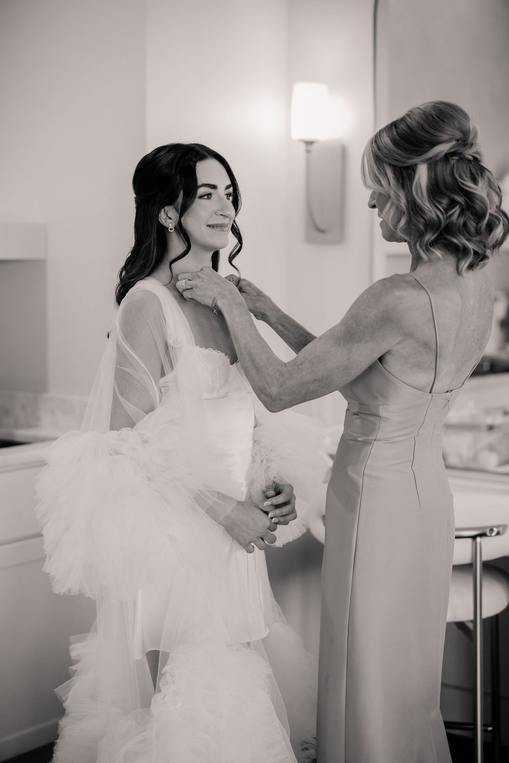 Black and white photo of bride being helped with necklace by bridesmaid during wedding preparations.