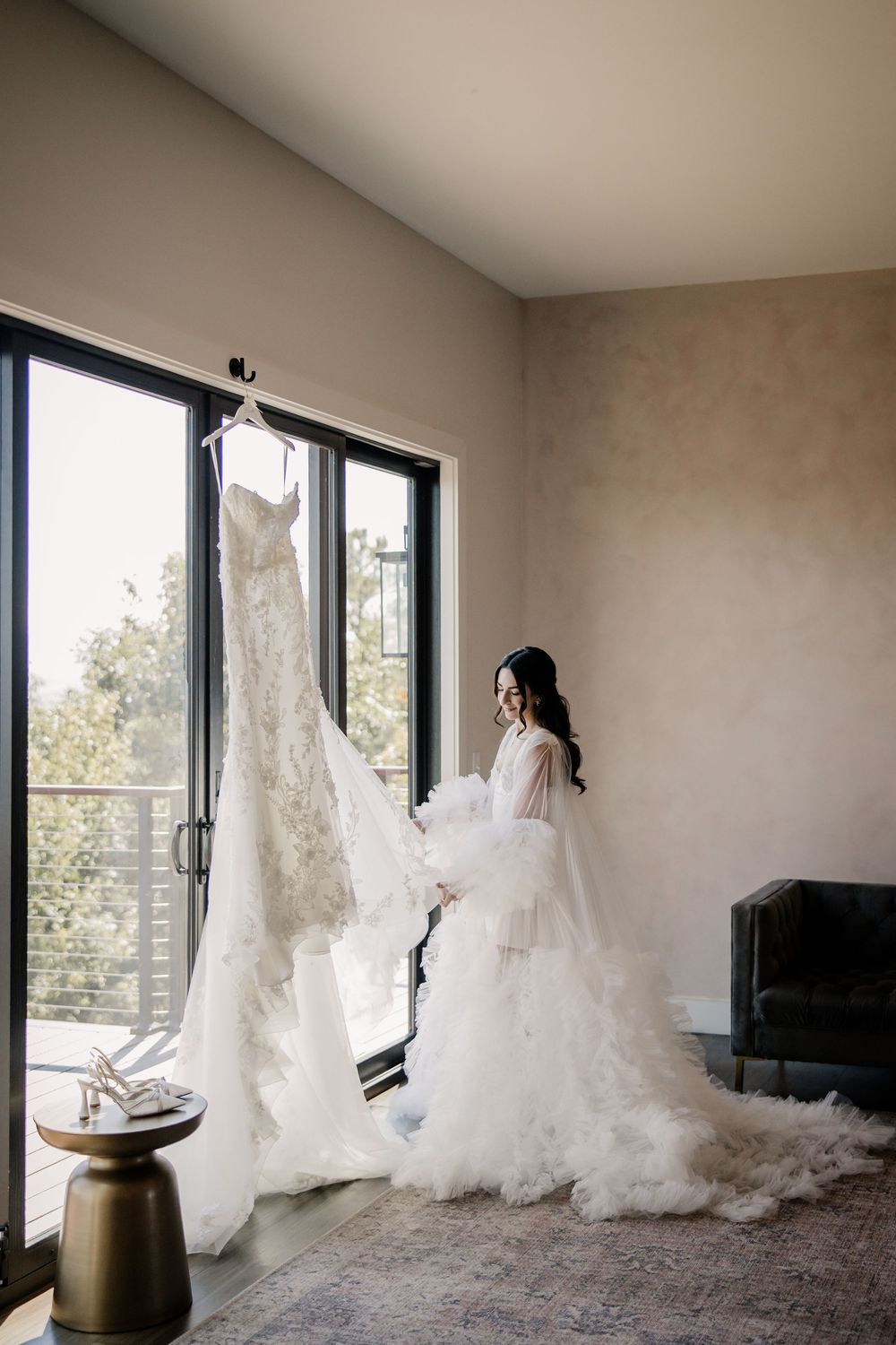 Bride in fluffy robe admires her lace wedding gown hanging by large window in bridal suite.