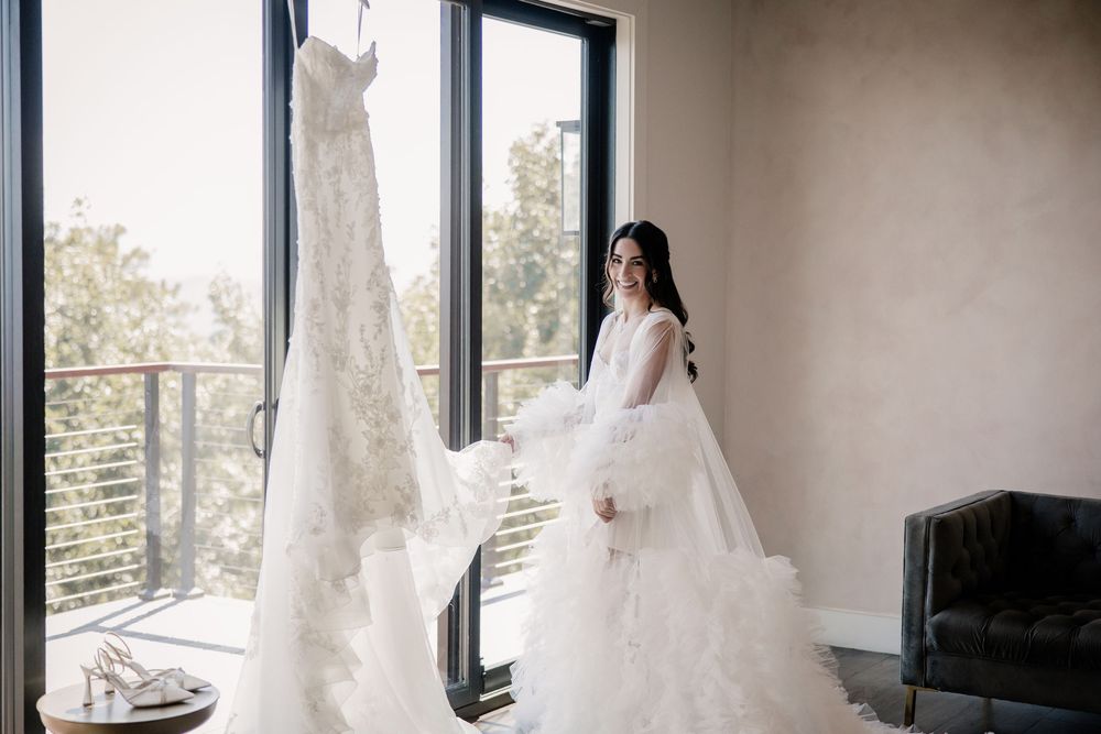 Bride in a fluffy white robe admires her lace wedding gown hanging by a large window in a bright room.
