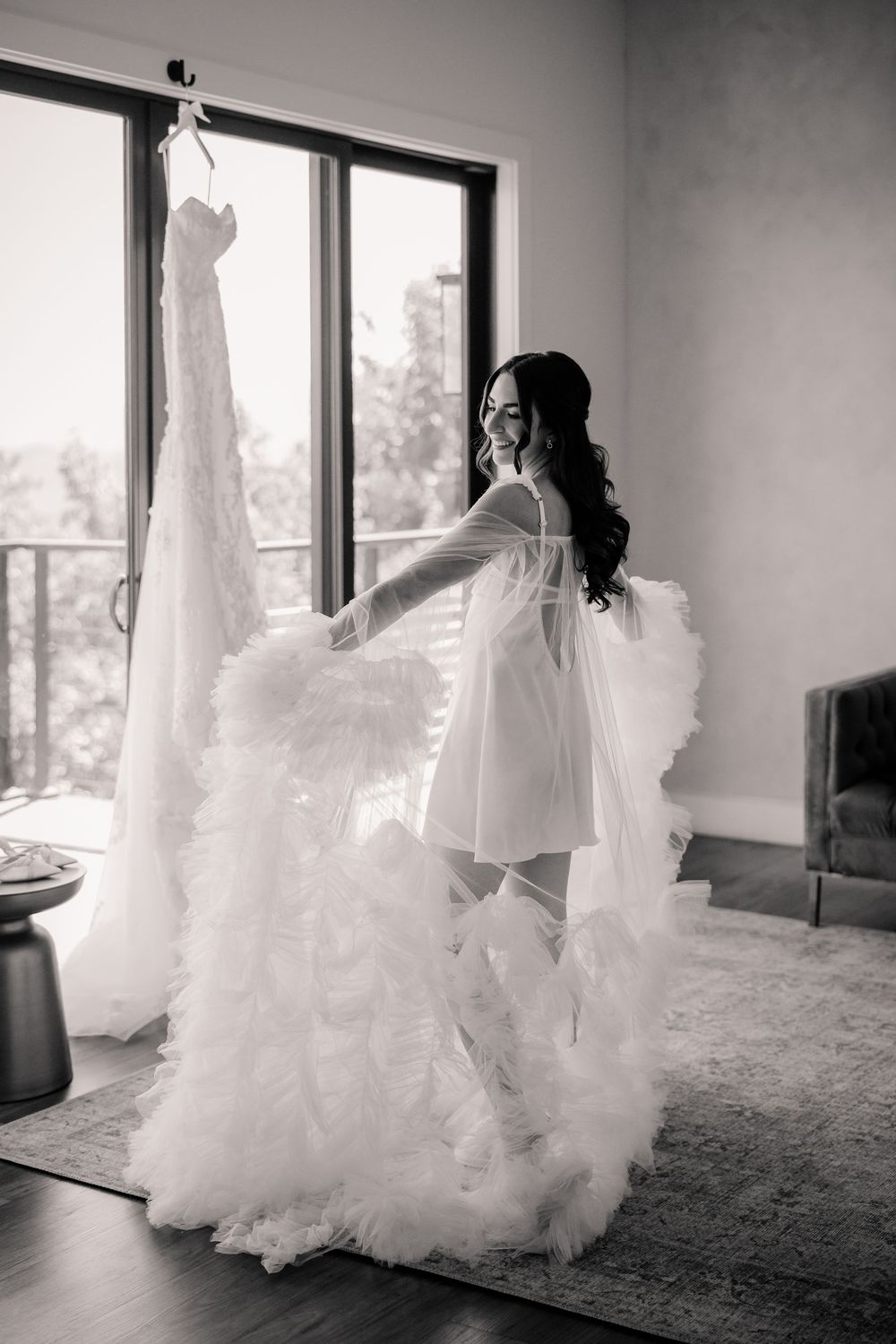 Bride in fluffy white robe smiling while holding her wedding gown in a black and white photo.