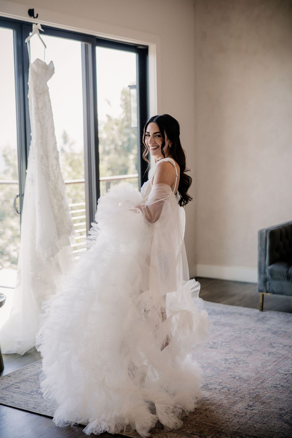Smiling bride in a fluffy white ruffled tulle gown holding her dress near a large window with her wedding dress hanging.
