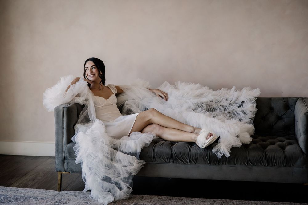 Bride in a short dress and fluffy robe reclines gracefully on a tufted gray velvet sofa.