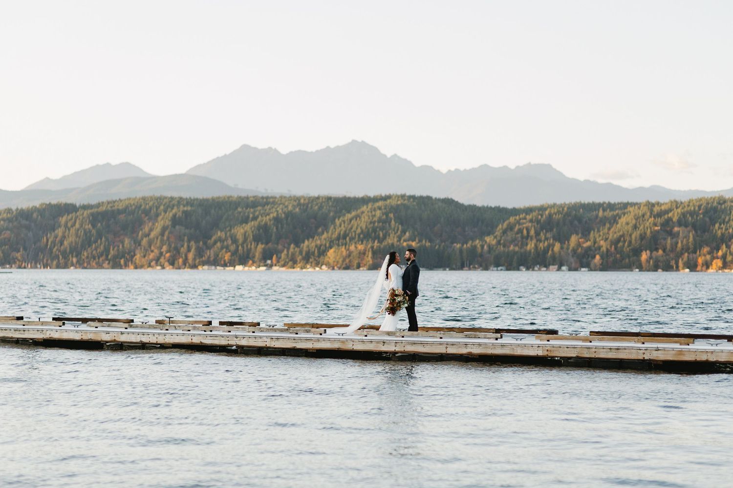 Bride and groom stand on a lakeside dock with mountain backdrop during fall wedding ceremony.