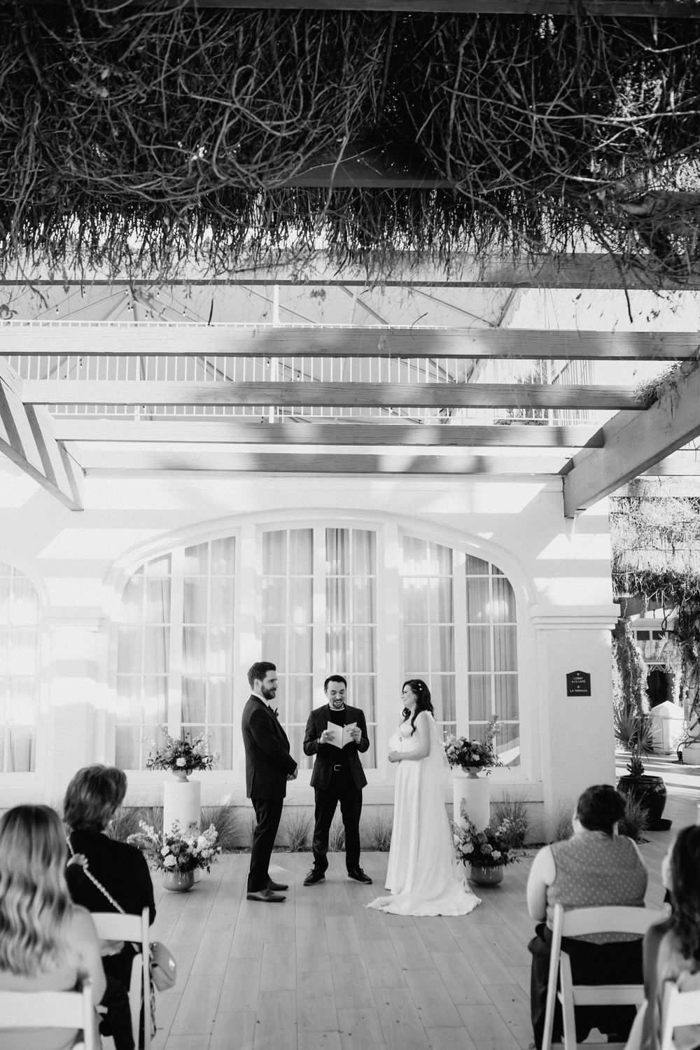 Black and white wedding ceremony photo with couple exchanging vows before officiant under pergola with seated guests.