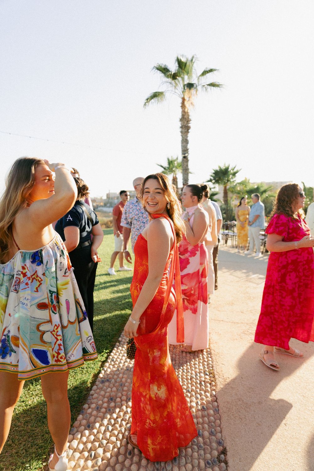 Woman in red tie-dye maxi dress smiling at outdoor garden party with palm trees and colorful dressed guests.