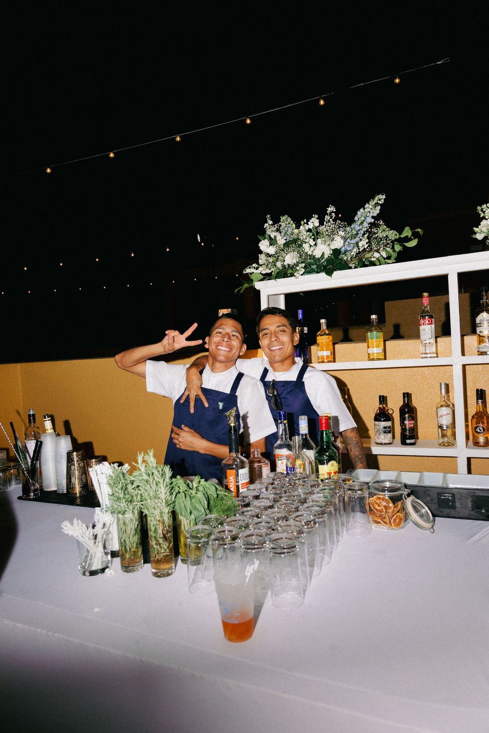 Two bartenders in blue aprons smile behind an outdoor bar with cocktails, glassware, and liquor bottles at a night event.