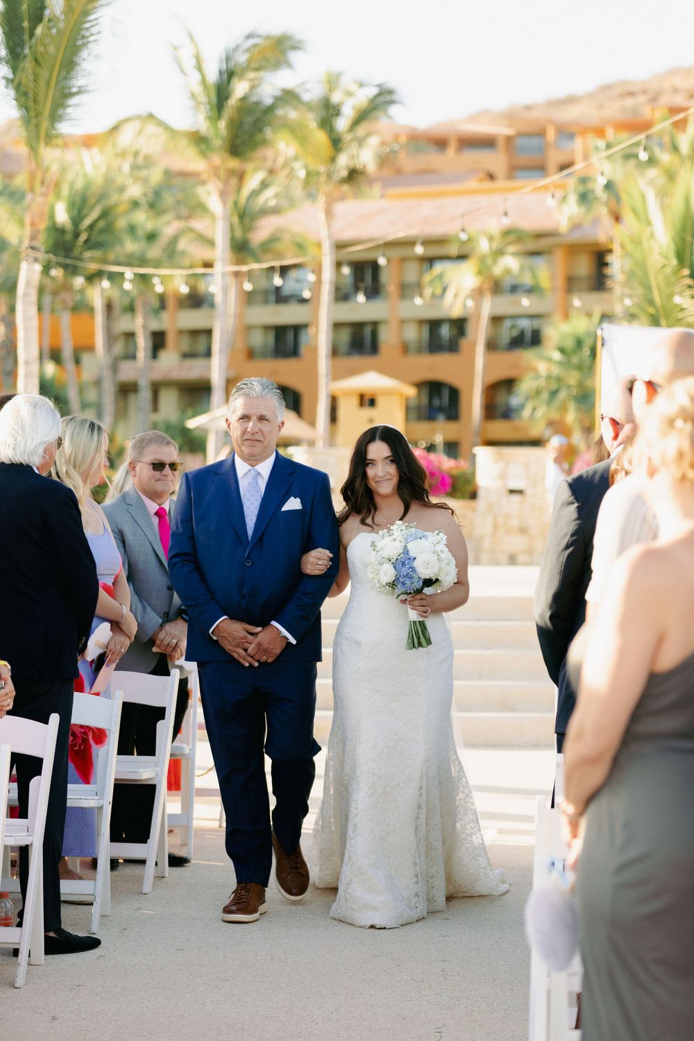 Bride in lace gown walks down outdoor aisle with escort at a tropical resort wedding ceremony.