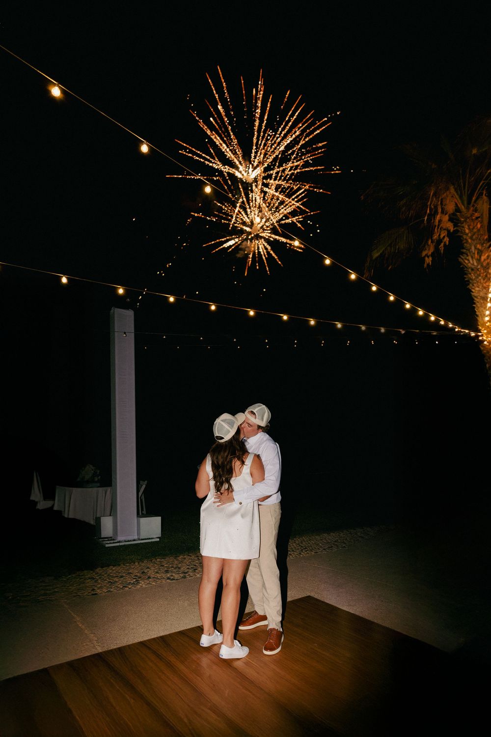 Couple embracing on dance floor under string lights with fireworks bursting in night sky at outdoor wedding reception.