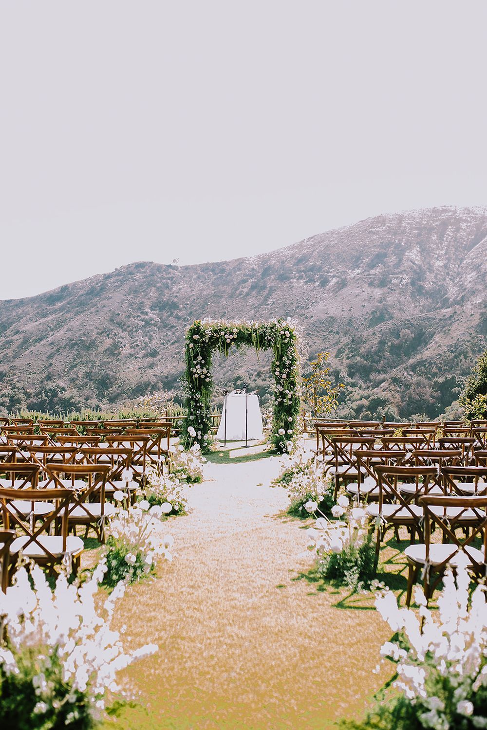 Rustic wooden benches and floral arrangements line an outdoor wedding ceremony space with mountain views in the background.