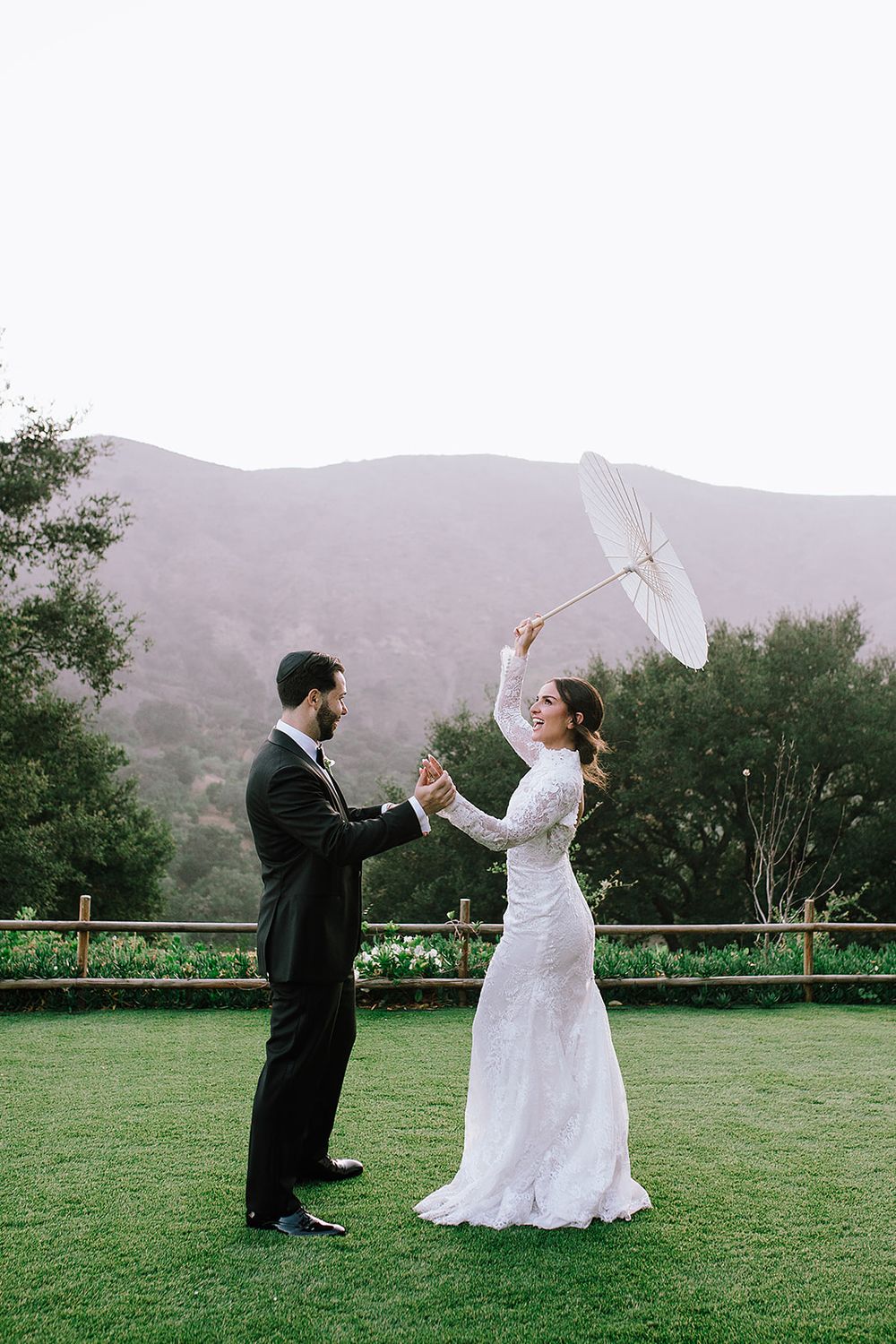 Playful couple having a sword fight with umbrellas on a grassy field with mountains in the background.