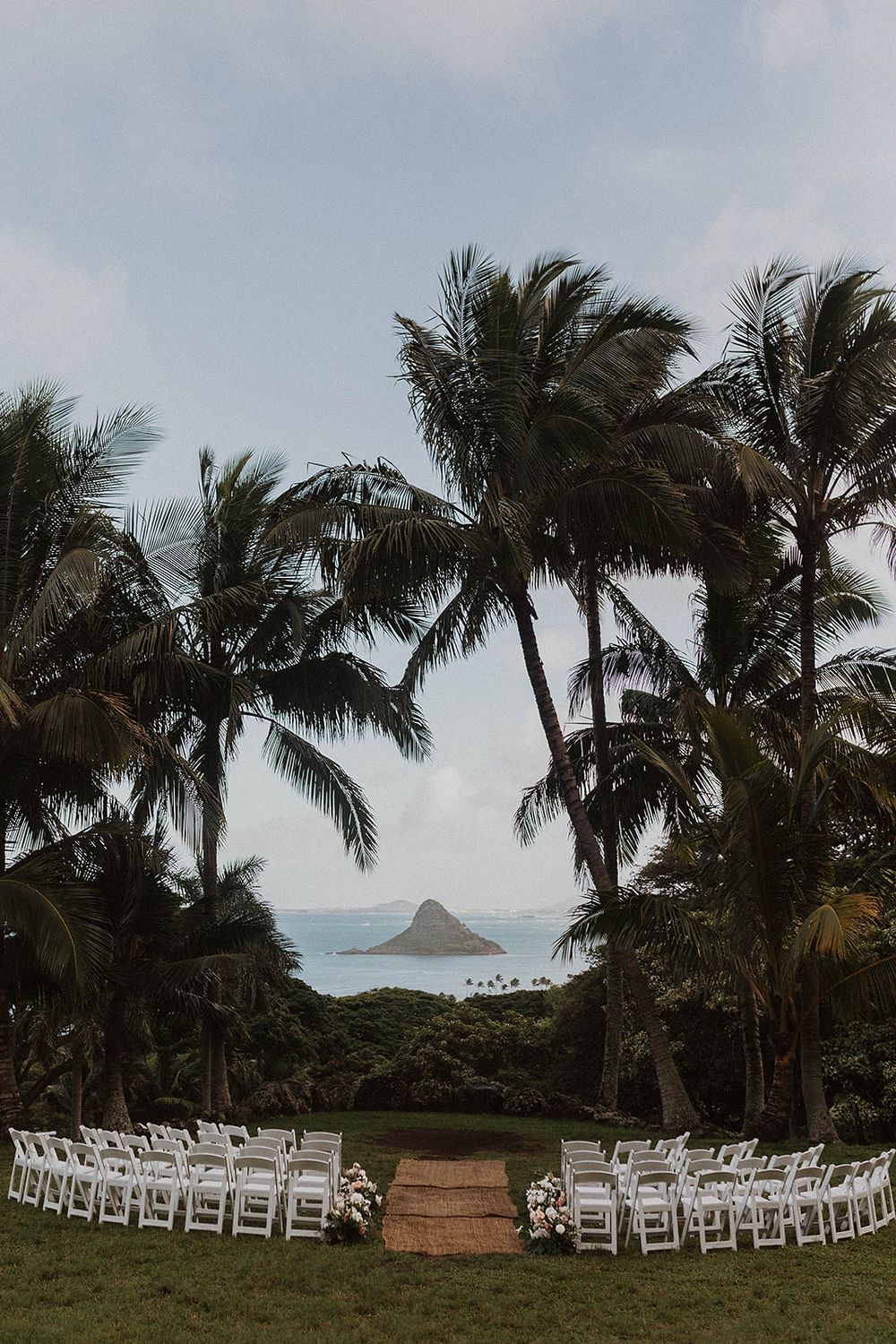 Outdoor wedding ceremony setup with white chairs on green lawn, framed by tall palm trees and ocean island views in Hawaii.