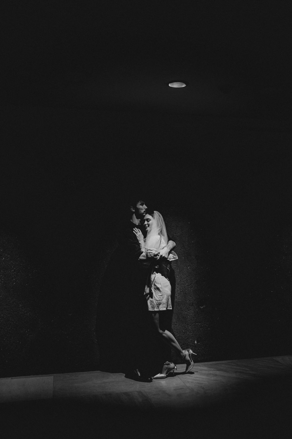 Black and white photo of a romantic couple embracing in a dimly lit hallway with dramatic shadows.