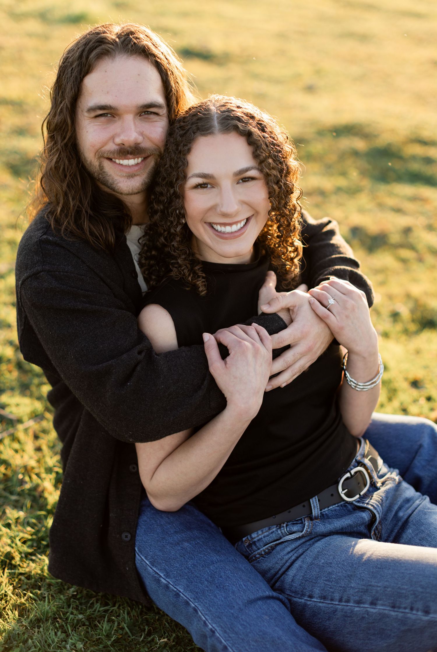 A couple embraces while sitting in a sunlit field wearing matching black sweaters and jeans during golden hour.