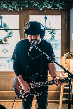 Man in black hat and headphones playing electric guitar and singing into microphone in cozy indoor setting.