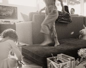 Black and white photo of a child jumping on a couch while another child draws on the floor in a living room.