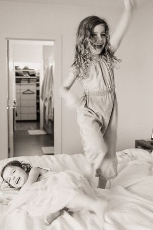 Two girls playing on a bed, one jumping joyfully with arms raised, the other lying down laughing, black and white photo.