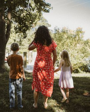 A woman in a red floral dress walks barefoot with two children through a sunny green park, holding a girl's hand.