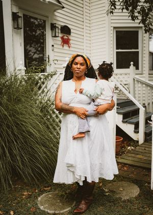 A Black woman in a white dress and brown boots holds a toddler while standing in front of a white house.