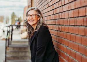 Smiling middle-aged woman with gray hair and glasses leaning against a red brick wall outdoors.