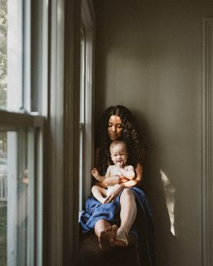 A mother with curly hair sits by a sunlit window holding her smiling baby wrapped in a blue towel.