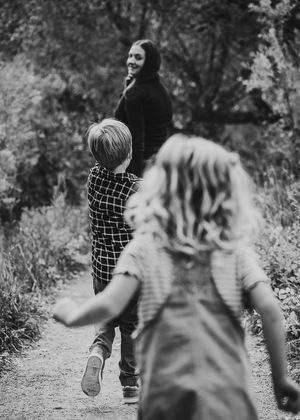 Black and white photo of two children running toward a smiling woman on a nature path.