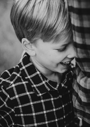 Black and white portrait of a smiling young boy in a plaid shirt, leaning against an adult.