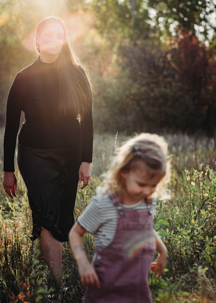 Woman in black dress walks behind toddler in pink overalls through sunlit meadow at golden hour.