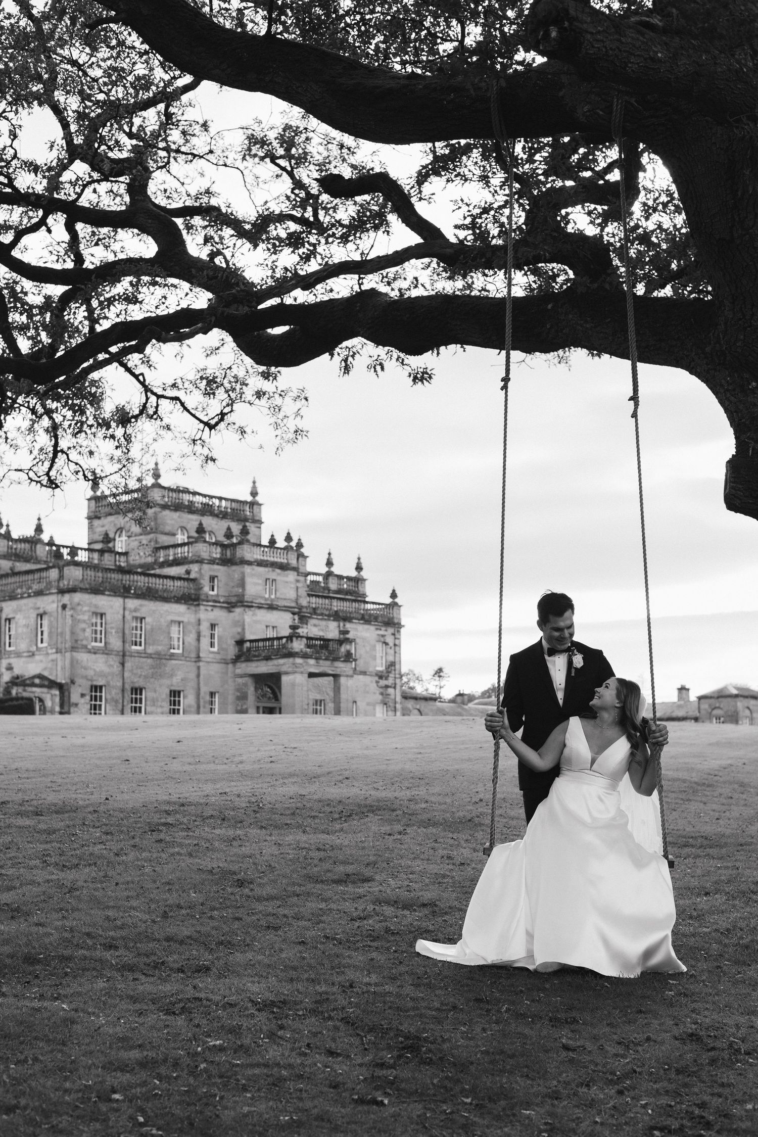 A couple on a tree swing poses against a grand estate mansion in a black and white wedding photograph.