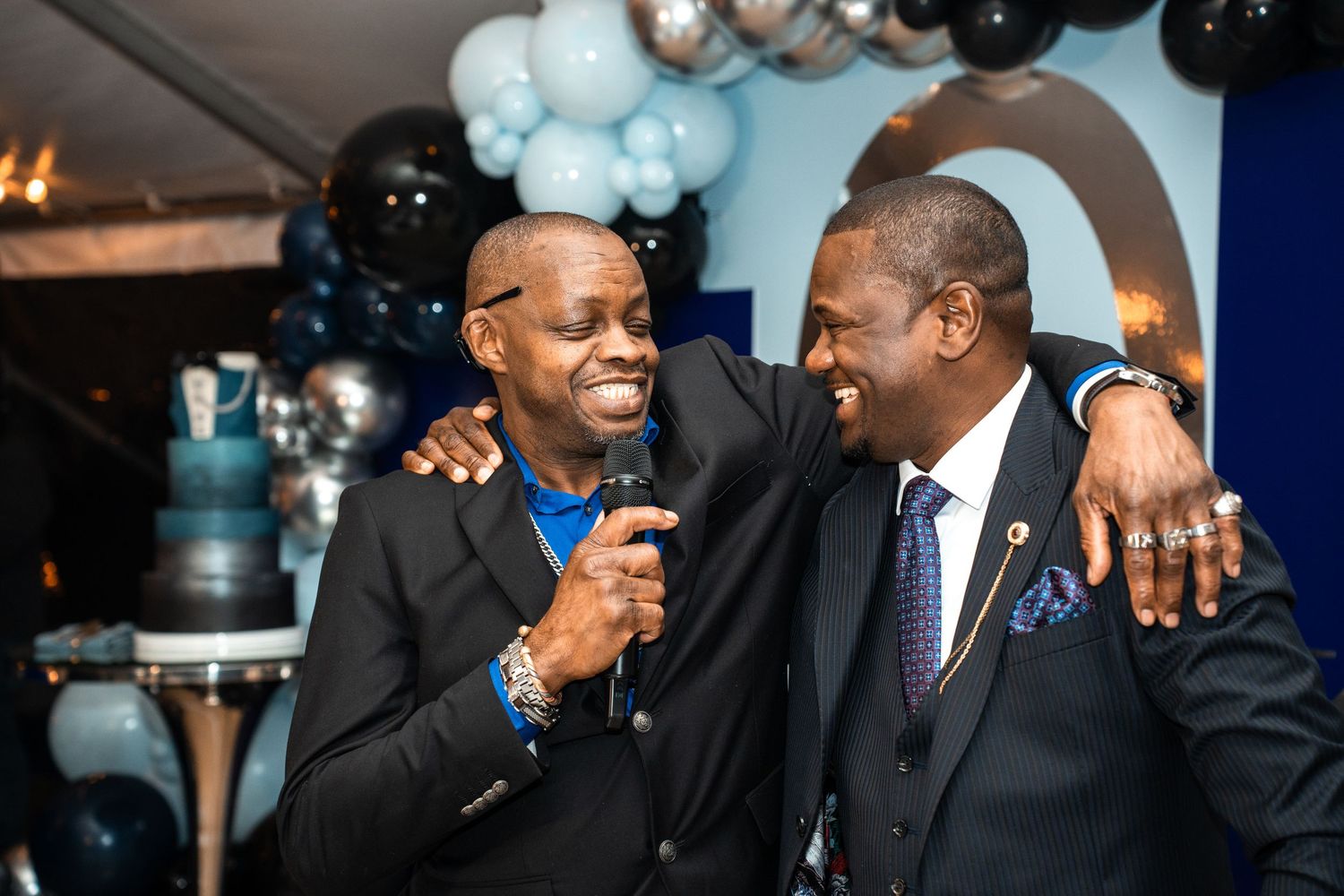 Two people sharing a joyful moment at an event with blue and black balloon decorations in the background.