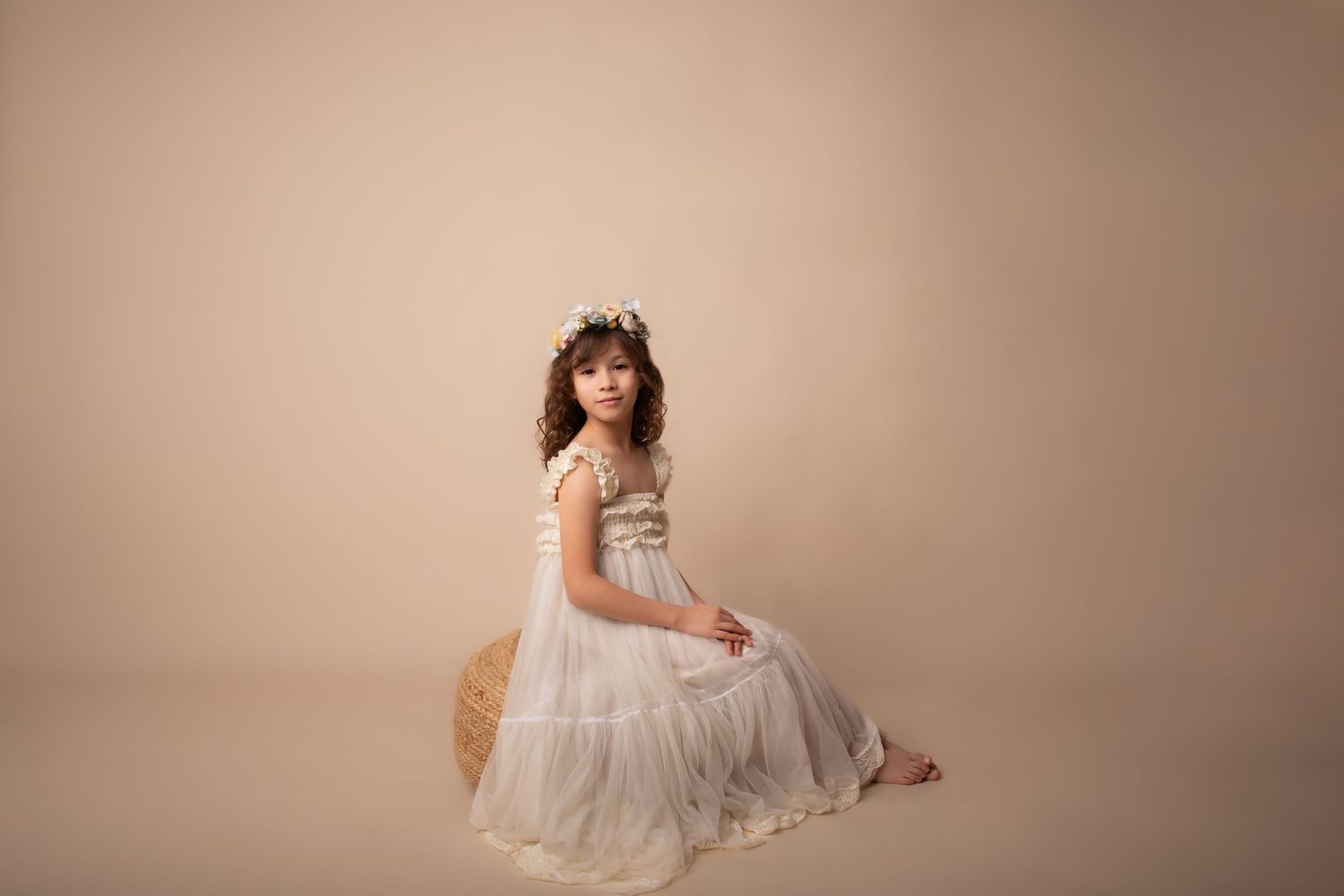 Ethereal portrait in flowing white dress on woven seat against neutral backdrop.