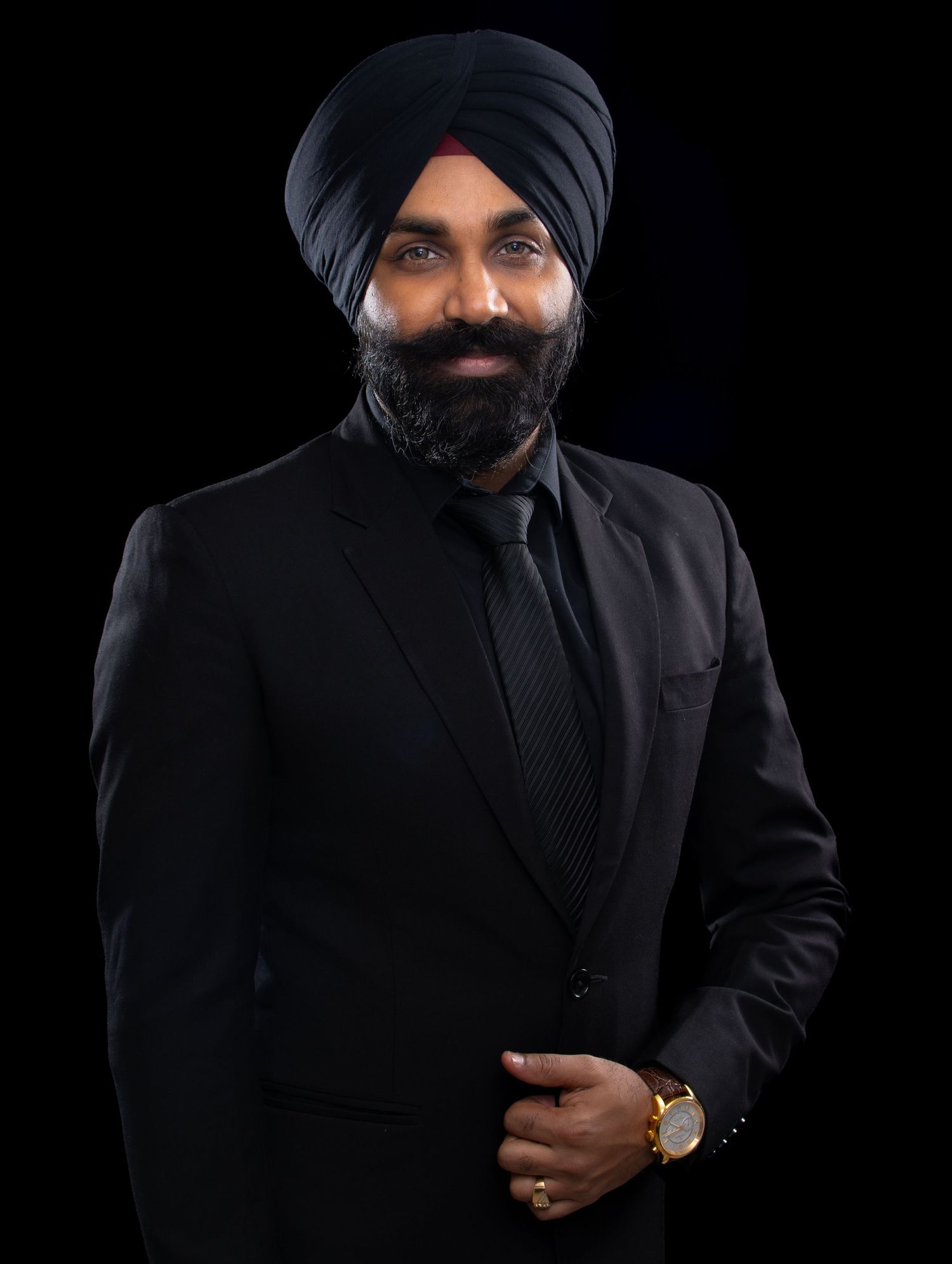 Professional portrait of a person in a black turban and suit with a black tie against a dark background.
