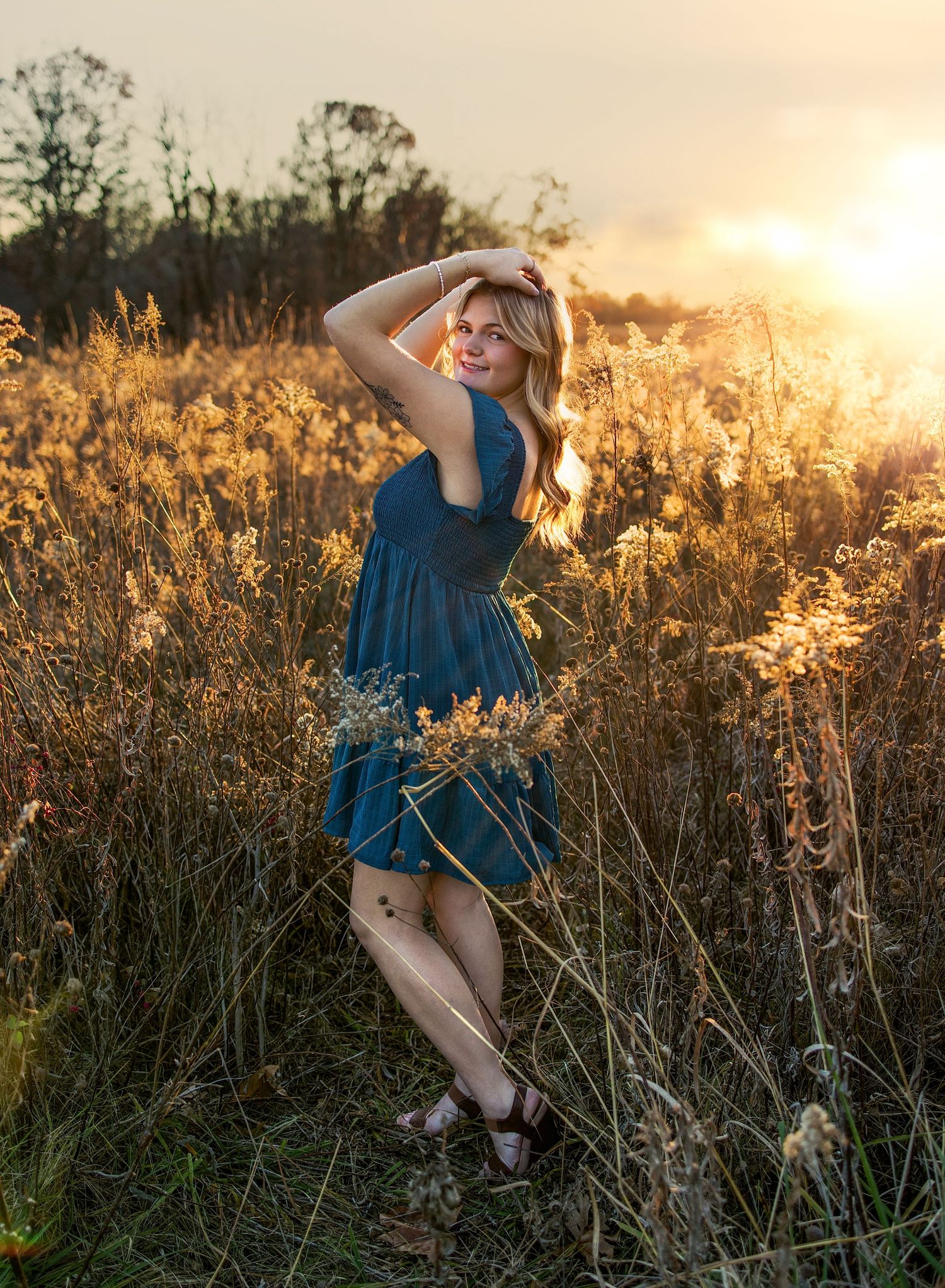 A senior girl in a blue dress sits gracefully among tall grasses at golden hour as sunlight creates a dreamy atmosphere.