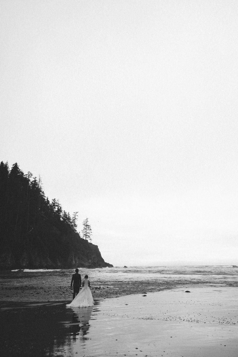 A couple walks along a misty Pacific Northwest beach with evergreen trees and cliffs in the background.