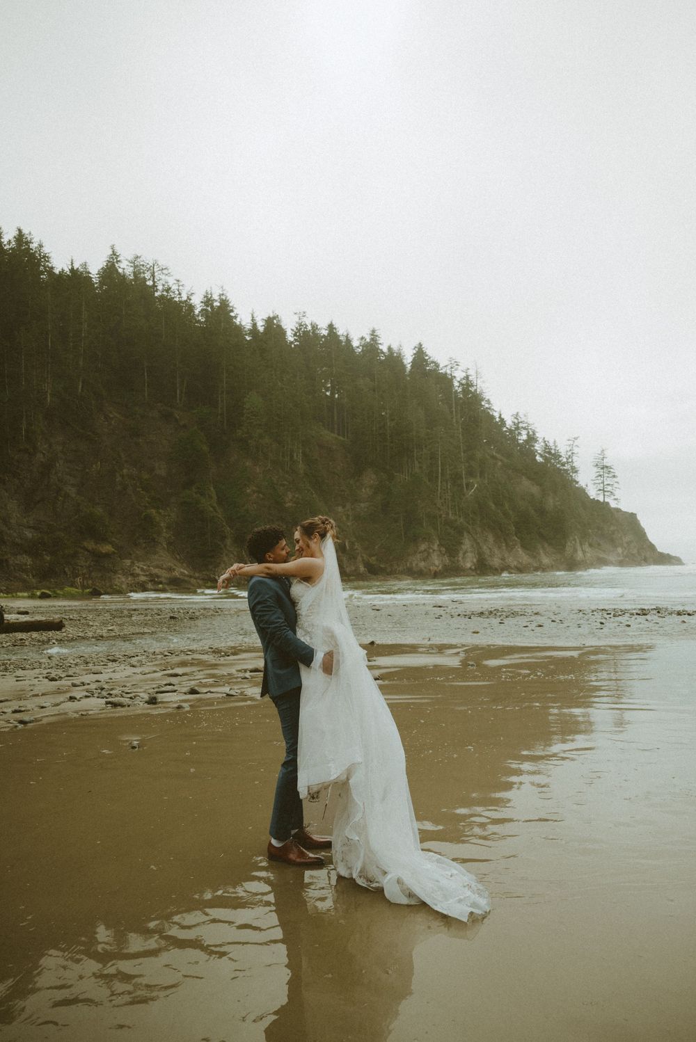 A couple in wedding attire shares an intimate moment on a misty Pacific Northwest beach with evergreen cliffs in background.