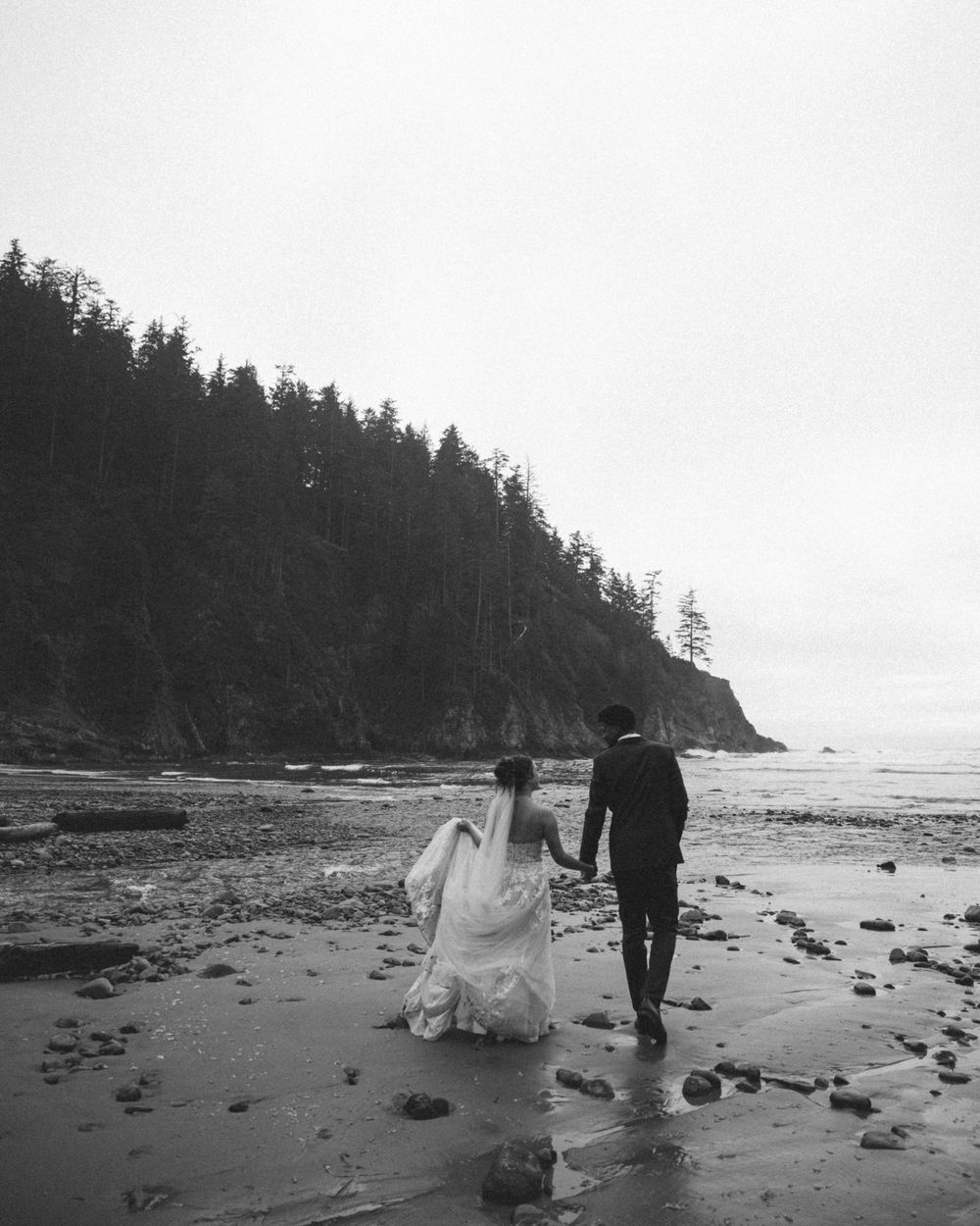A couple walks hand in hand along a misty Pacific Northwest beach with evergreen trees lining the rocky shore.