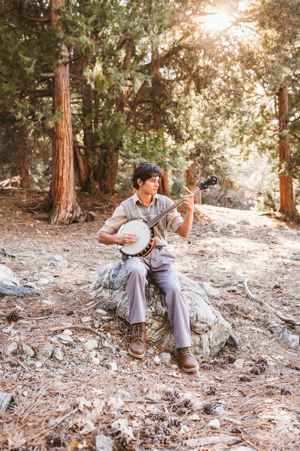 A musician plays banjo while sitting on a rock in a sunlit forest setting with redwood trees in the background.