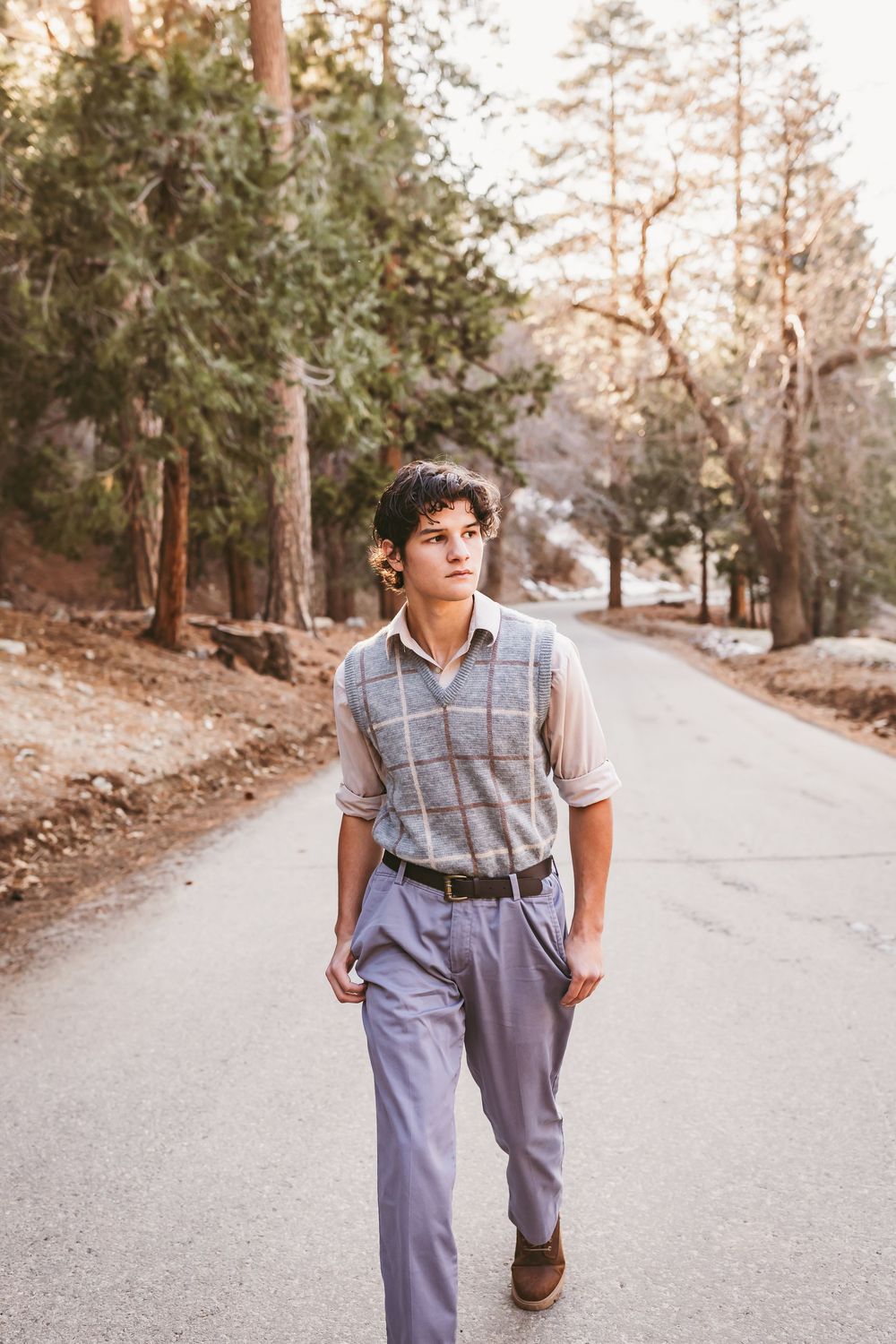 A person in vintage-style gray trousers and plaid vest walks along a scenic mountain road on a sunny afternoon.