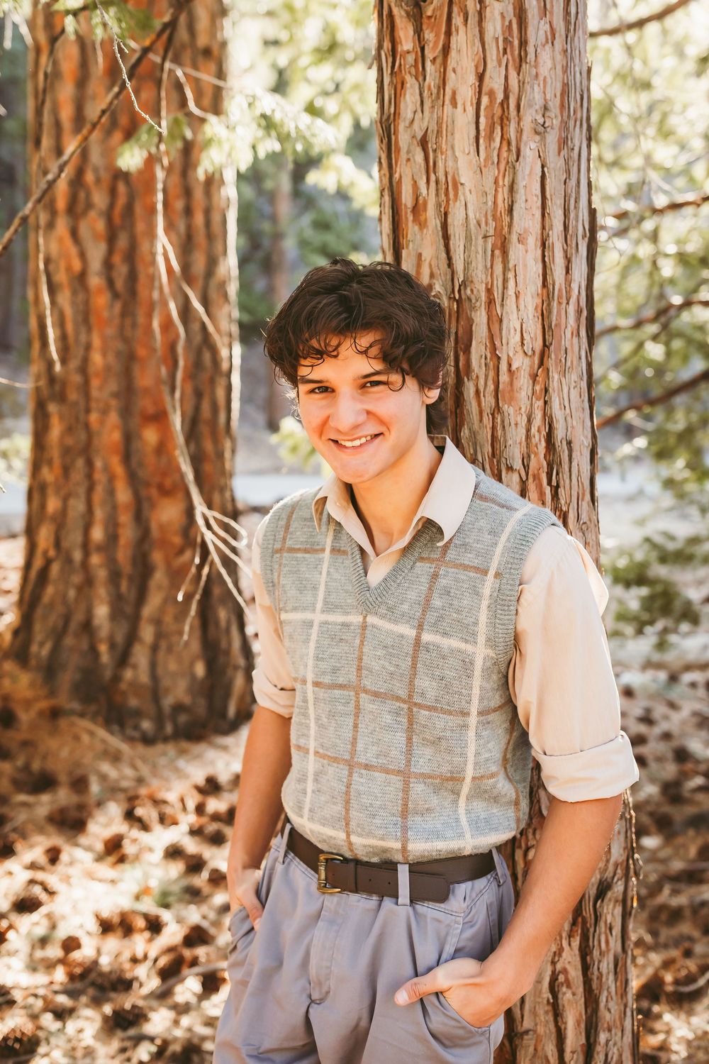 A person in a grey sweater vest and white shirt stands casually against a redwood tree trunk in dappled sunlight.