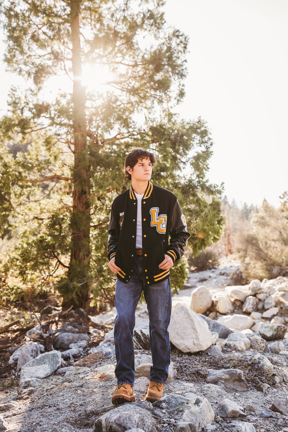 A letterman jacket-clad figure stands on rocky terrain at sunset with sunlight streaming through pine trees in the background.