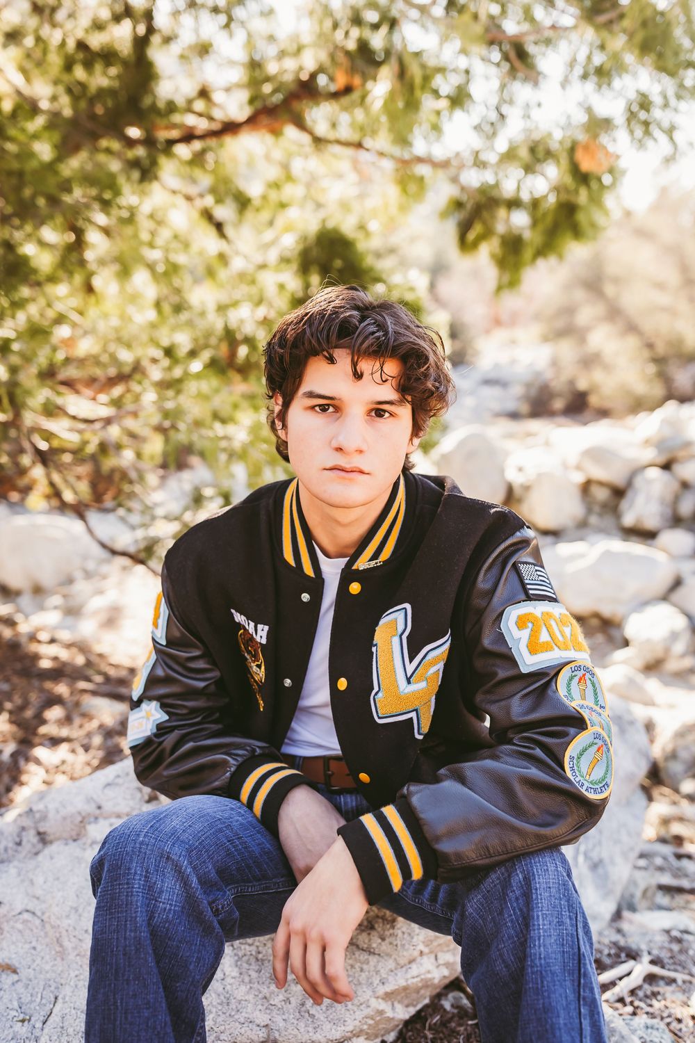A varsity letterman jacket with patches is worn while sitting on rocks in a natural outdoor setting.