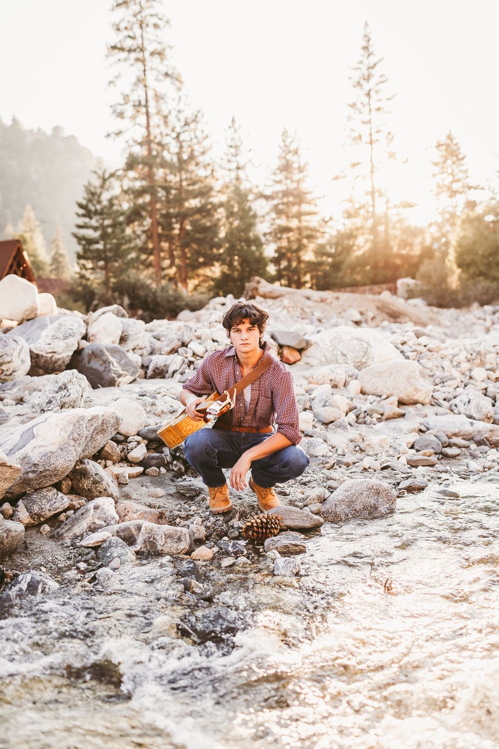 Person in casual attire sits among rocky terrain during golden sunset with evergreen trees in background.