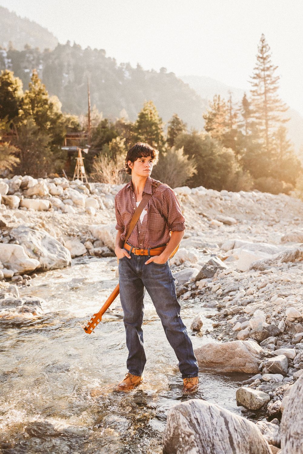 Person in plaid shirt and jeans explores rocky mountain landscape at sunset with pine trees in distance.