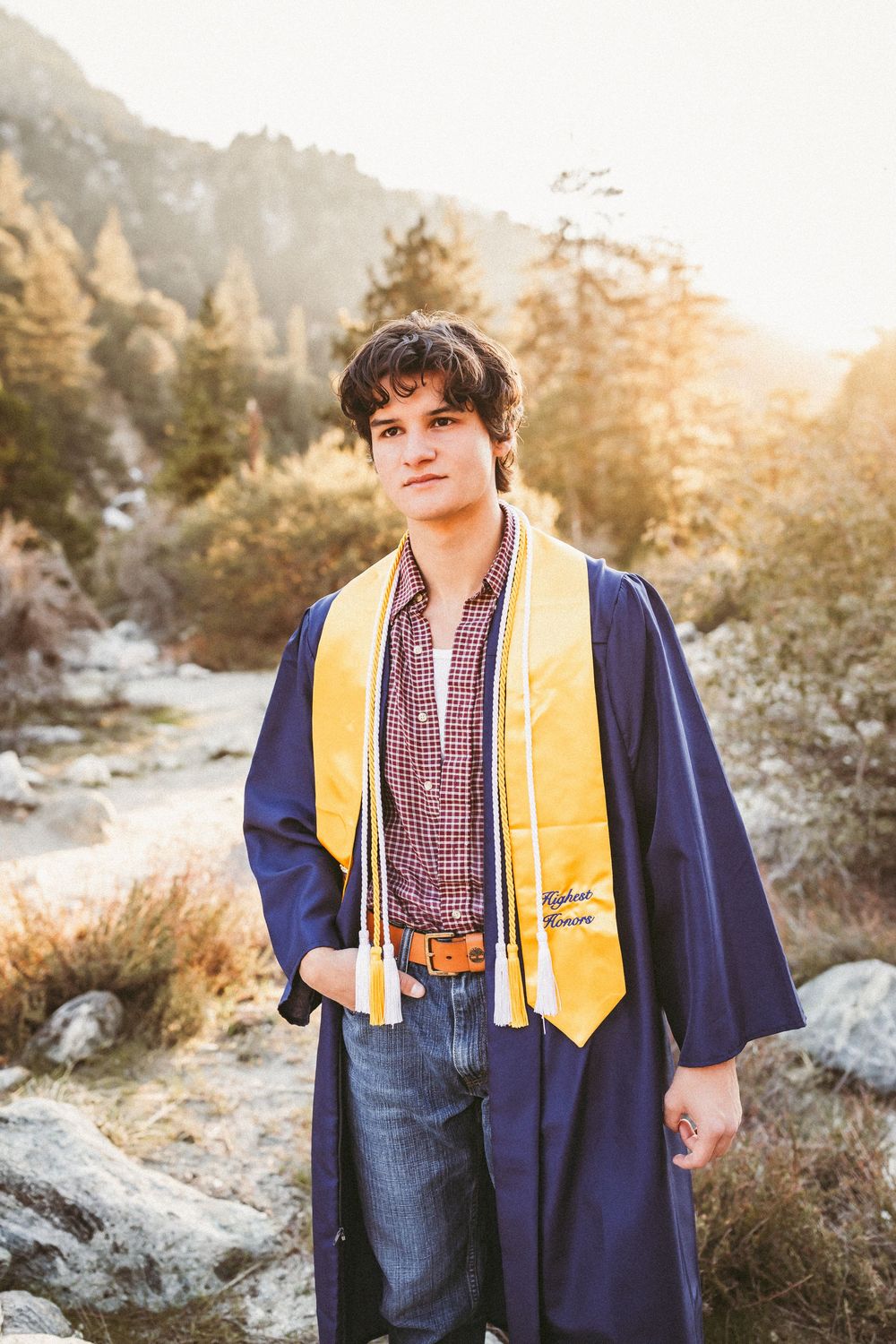 A graduate in a navy cap and gown with yellow stole poses outdoors against a sunlit mountain landscape.