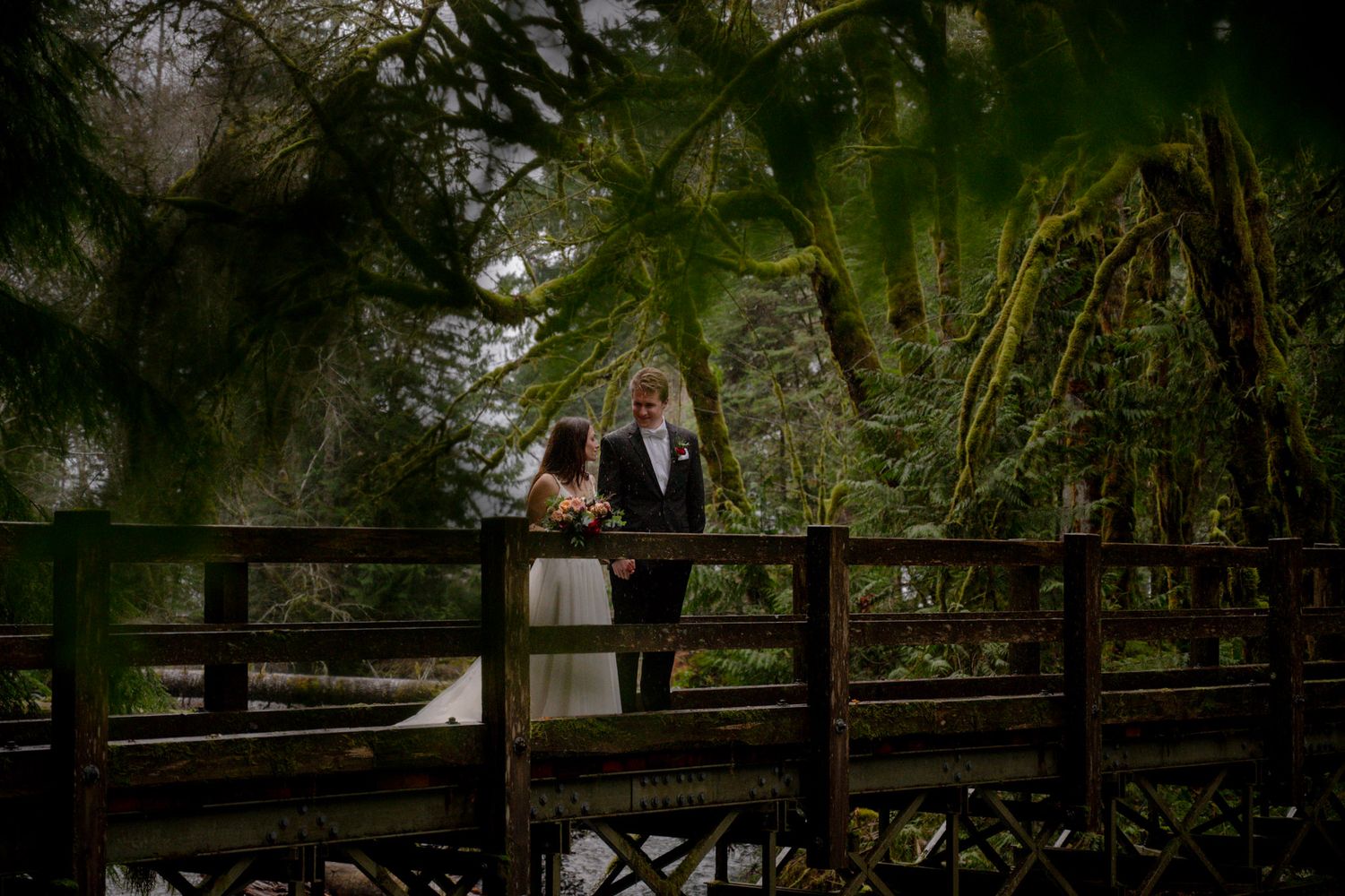A bride and groom share a romantic moment on a wooden bridge surrounded by lush green forest.