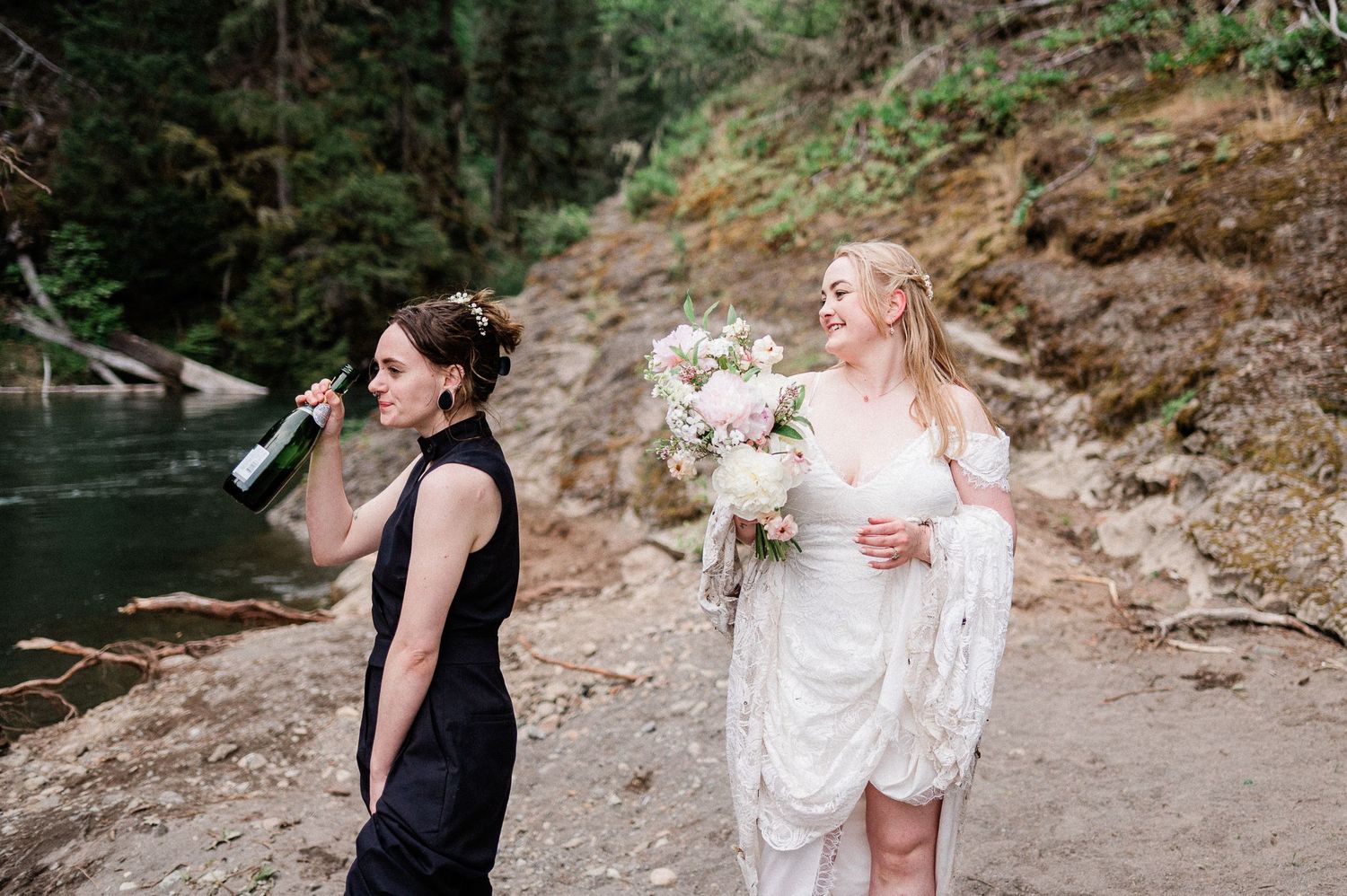 Two brides share a laugh by a scenic forest river with rocky cliffs in the background.