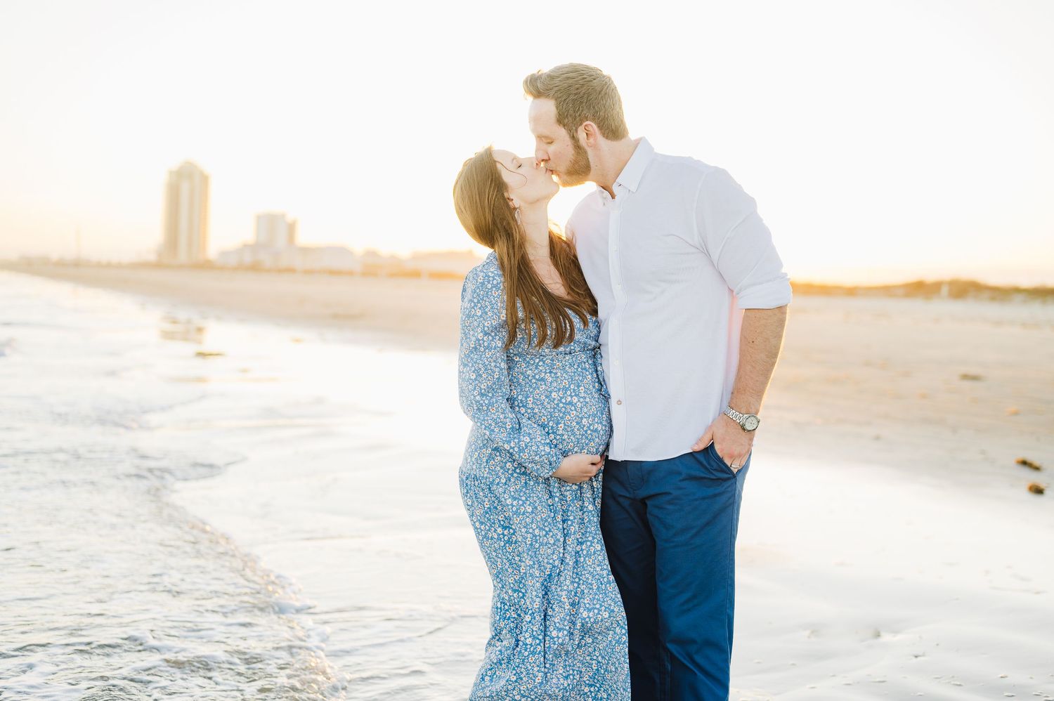 A couple stands together on a sandy beach during golden hour as waves gently roll in behind them.