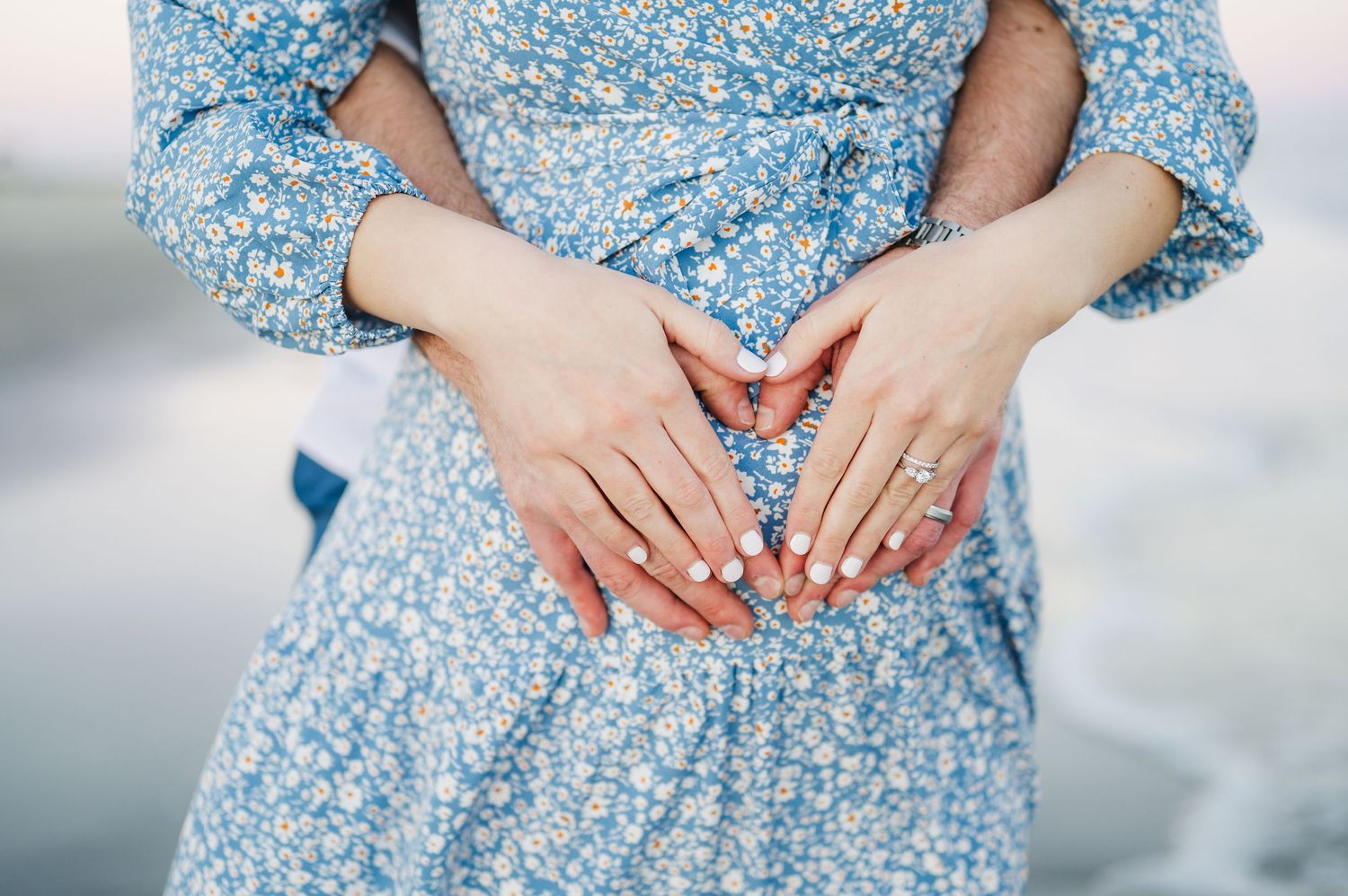 Hands with blue nail polish rest on a light blue floral dress against a soft blurred background.